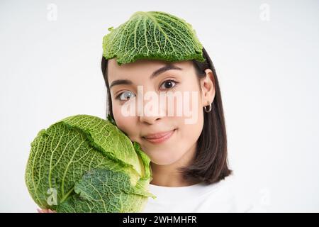 Primo piano ritratto di bella e sana donna vegana, tenendo lattuga, ha cavolo piombo sulla testa, sfondo bianco Foto Stock