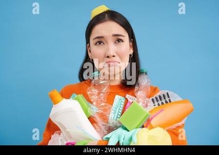 Ritratto di ragazza piangente triste con rifiuti di plastica, raccoglie rifiuti di riciclaggio, sembra miserabile, sfondo blu Foto Stock