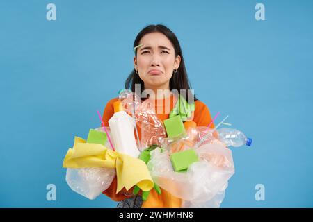 Piangendo femmina asiatica che tiene rifiuti di plastica, spazzatura nelle mani e guardando sconvolto, deve riciclare, sfondo blu Foto Stock