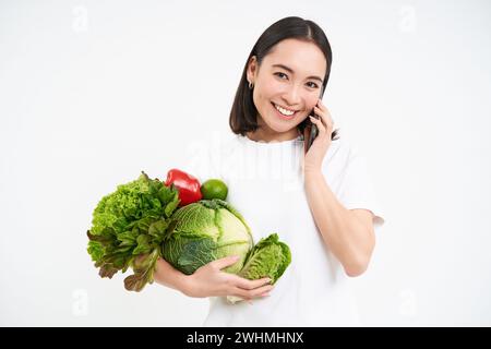 Donna coreana sorridente, parla al telefono cellulare, ordina verdure, tiene verde biologico, cibo crudo, isolato su sfondo bianco Foto Stock