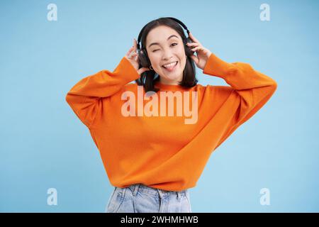 Felice donna cinese in cuffie, ascolta la musica, ama la canzone preferita nella sua playlist, si trova su sfondo blu Foto Stock