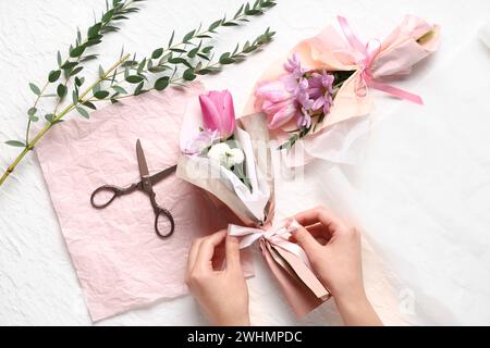 Mani femminili con piccoli mazzi di splendidi fiori primaverili in carta da imballaggio, eucalipto e forbici su sfondo bianco. Giornata internazionale della donna Foto Stock