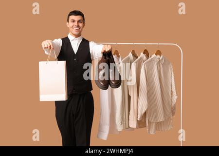 Bel giovane uomo felice che tiene in mano borsa, stivali e scaffale con camicie pulite su sfondo marrone Foto Stock