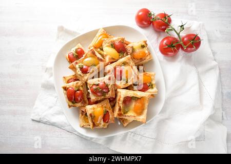 Spuntini di pasta sfoglia al forno con formaggio spalmabile, pomodori e pesto alle erbe, finger food da festa su un piatto bianco, vista dall'alto da ab Foto Stock