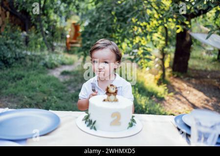 Il bambino con i capelli biondi ondulati e la camicia bianca si siede al tavolo vicino alla torta di compleanno all'aperto. Foto Stock