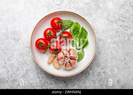 Cucina italiana con pomodori ciliegini, basilico verde fresco e spicchi d'aglio, vista dall'alto sul piatto su fondo di pietra bianca Foto Stock