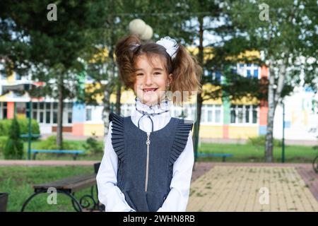 Allegra ragazza divertente con un sorriso senza denti in uniforme scolastica con archi bianchi nel cortile scolastico. Ritorno a scuola, 1° settembre. Un ettaro Foto Stock