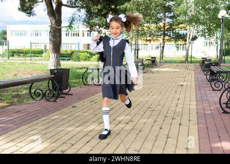 Allegra ragazza divertente con un sorriso senza denti in uniforme scolastica con archi bianchi che corrono nel cortile scolastico. Ritorno a scuola, 1° settembre. Foto Stock