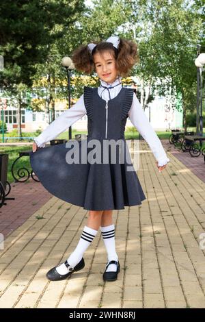 Allegra ragazza divertente con un sorriso senza denti in uniforme scolastica con archi bianchi nel cortile scolastico. Ritorno a scuola, 1° settembre. Un ettaro Foto Stock