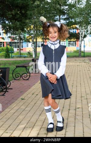 Allegra ragazza divertente con un sorriso senza denti in uniforme scolastica con archi bianchi nel cortile scolastico. Ritorno a scuola, 1° settembre. Un ettaro Foto Stock