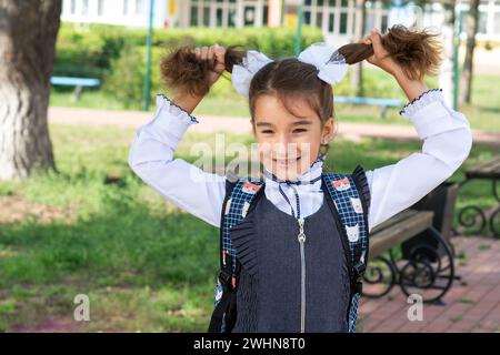 Allegra ragazza divertente con un sorriso senza denti in uniforme scolastica con archi bianchi nel cortile scolastico. Ritorno a scuola, 1° settembre. Happ Foto Stock