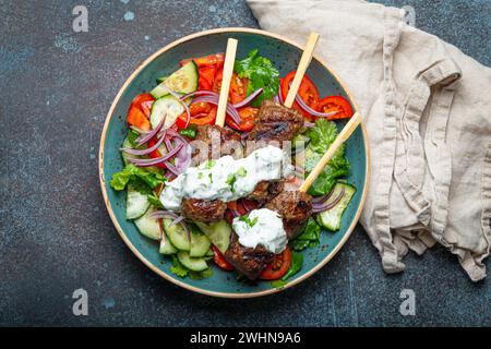 Spiedini grigliati di carne di manzo spiedini su bastoncini serviti con insalata di verdure fresche su piatto su sfondo rustico di cemento dall'alto. Foto Stock