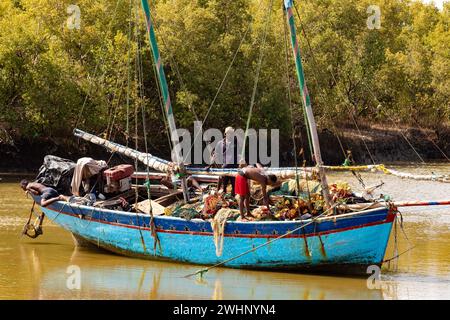 Due uomini riparano una rete da pesca su una barca blu ormeggiata sulla riva del fiume Morondava in Madagascar Foto Stock