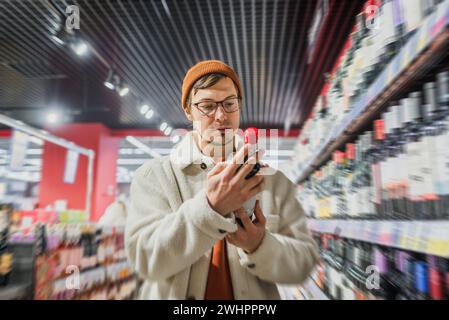 Un uomo sceglie una bottiglia di vino in un supermercato nel reparto alcolici. Un giovane uomo concentrato con un berretto all'arancia e un cappotto di panna alla moda esamina attentamente una bottiglia di vino in un corridoio di supermercato ben illuminato e colorato, con scaffali riforniti con una vasta gamma di prodotti. Foto Stock
