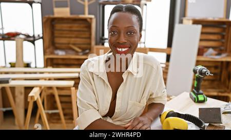 Donna afroamericana sorridente falegname seduto con fiducia al tavolo della falegnameria nel suo impegnato laboratorio al coperto Foto Stock