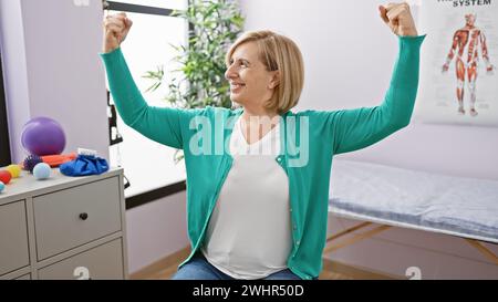 Una donna bionda sorridente di mezza età che si flette i muscoli in una stanza clinica di riabilitazione, mostrando forza e positività. Foto Stock