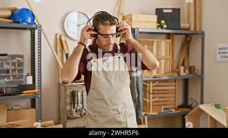 Uomo ispanico in officina che indossa occhiali di sicurezza, circondato da utensili per la lavorazione del legno e banco da lavoro. Foto Stock