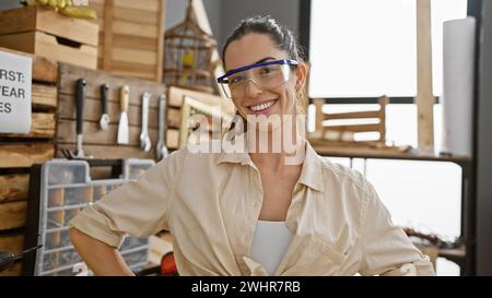 Bellissima giovane donna ispanica, un falegname sorridente, orgogliosa della sua officina di falegnameria, che indossa occhiali di sicurezza Foto Stock