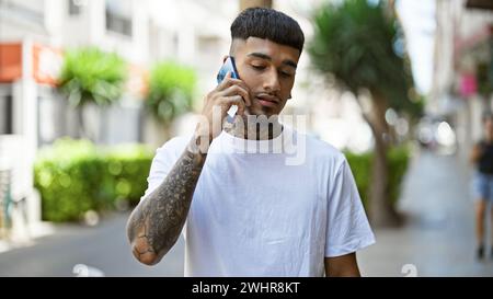 Giovane latino tatuato e simpatico, coinvolto seriamente in una conversazione telefonica, in piedi all'aperto sulla strada della città illuminata dal sole, trasuda vibrazioni rilassate mentre si usa la Foto Stock