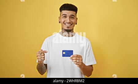 Giovane latino sicuro di sé, sorridente allegramente, che punta alla sua carta di credito, isolato su un vibrante sfondo giallo. espressione finanziaria positiva di Foto Stock