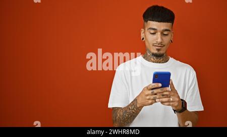 Giovane latinoamericano tatuato, avvolto nel suo smartphone su sfondo rosso isolato, ritratto di un singolo immer dall'aspetto serio e concentrato Foto Stock