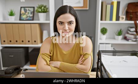 Giovane donna ispanica sicura di sé in un ufficio moderno con le braccia incrociate Foto Stock