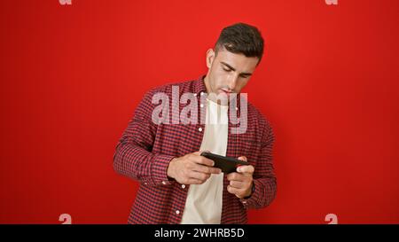 Un giovane ispanico molto attraente, immerso in un videogioco, isolato su uno sfondo rosso, che ascolta la musica dal suo telefono, gioca Foto Stock