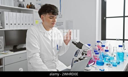 Un giovane con un camice da laboratorio bianco esamina una penna mentre lavora con un microscopio in un laboratorio ben attrezzato. Foto Stock