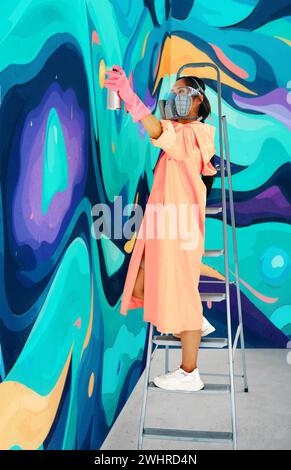 Street artist donna in maschera respiratoria che dipinge graffiti colorati su una parete in piedi su una scala Foto Stock