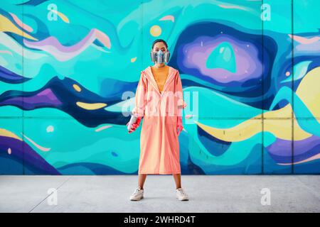 Ritratto a figura intera di una Street artist donna in maschera respiratoria in piedi vicino al muro dei graffiti Foto Stock
