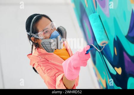 Street artist donna in maschera respiratoria che dipinge graffiti colorati con rullo per pittura sulla parete Foto Stock