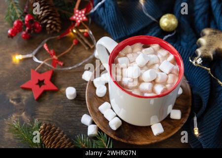 Accogliente tazza di cioccolata calda natalizia con marshmallow e luci natalizie, maglione lavorato a maglia su sfondo di legno. Foto Stock