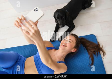 Ritratto di una giovane donna che si forma in forma, sdraiata sul tappetino e scattata selfie con un cane in salotto, allenati con il suo animale domestico Foto Stock