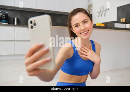 Una bella ragazza sorridente registra video mentre fa sport a casa, guarda lo smartphone, scatta selfie sul telefono cellulare, allena indo Foto Stock