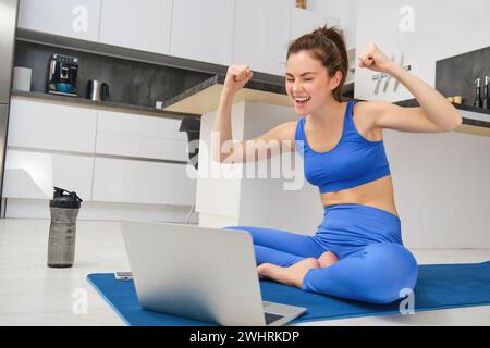 Ritratto di una giovane donna eccitata, seduta con un laptop sul tappetino, che fa il tifo per il cliente, che dà lezioni online di yoga, lezioni di fitness a distanza, W Foto Stock