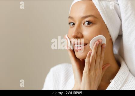 La bella donna con l'abito e l'asciugamano sulla testa pulisce la pelle del suo viso con tamponi di cotone. Pulizia e cura sana del corpo dopo sh Foto Stock