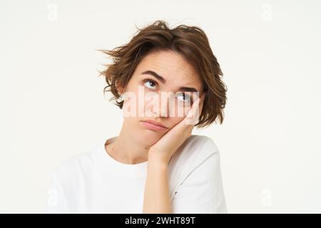 Primo piano di una giovane donna annoiata, tira gli occhi, si appoggia alla mano e sembra riluttante e sconvolta, isolata su sfondo bianco Foto Stock