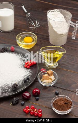 Ingredienti per la cottura della torta di bacche. Farina in piatto nero, cacao in polvere. Dosatore con farina, bicchiere di latte, uova rotte e sale, frusta di metallo sulla linguetta Foto Stock