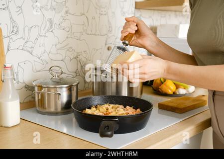 Vista ravvicinata delle mani femminili che grattugia il parmigiano in una padella con pasta cotta e crema di funghi Foto Stock