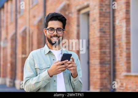 Un uomo arabo informale guarda la fotocamera con un ampio sorriso mentre usa il cellulare sullo sfondo di un edificio in mattoni. Utente disattento di gadget in occhiali classici divertiti a giocare con la nuova applicazione mobile online. Foto Stock