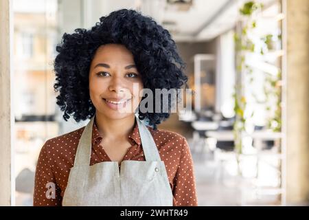 Ritratto di una donna sorridente che indossa il grembiule sopra la camicetta mentre sorride alla fotocamera su sfondo sfocato. Cameriera ottimista che accoglie i clienti in un ristorante vuoto durante il giorno. Foto Stock