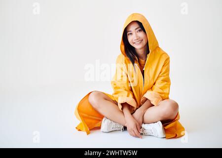 Sorridente ragazza asiatica con bretelle vestita con un impermeabile giallo brillante che posa con cappuccio sulla testa seduta sul pavimento Foto Stock