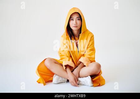 Sorridente ragazza asiatica con bretelle vestita con un impermeabile giallo brillante che posa con cappuccio sulla testa seduta sul pavimento Foto Stock