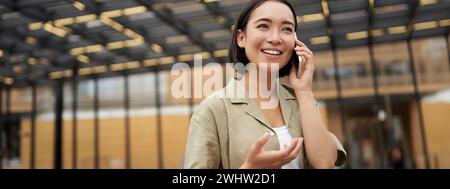 Giovane donna asiatica sorride, parla di cellulare e passeggiate nel centro della città, pone all'aperto Foto Stock