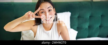 Positiva donna asiatica sdraiata a letto, mostrando segno di pace, gode felice mattina, svegliandosi ottimista, rimanendo nella sua camera da letto Foto Stock