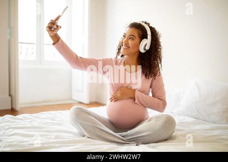 Donna incinta di colore brillante con cuffie che ascolta musica e scatta selfie Foto Stock