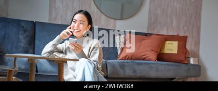 Foto verticale di un'elegante donna coreana seduta sul pavimento di casa, utilizzando il telefono cellulare, tenendo in mano lo smartphone e sorridendo Foto Stock