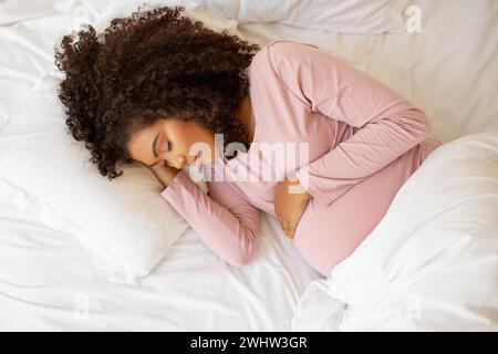 Bella e tranquilla donna incinta di colore che riposa sul letto bianco di casa Foto Stock