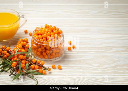 Spina di mare tè caldo sano in tazza di vetro trasparente, bacche mature in vaso di vetro e rami con foglie su fondo rustico di legno bianco, per la pelle Foto Stock