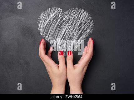 Un cuore disegnato con gesso bianco su una tavola nera e due mani femminili Foto Stock